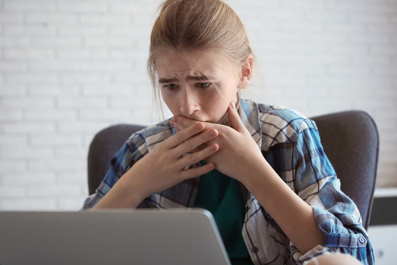 A shocked, worried young girl looks at a computer with her hand over her mouth